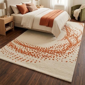 Puede incluir: Una alfombra de color crema con un patrón de puntos en relieve de color óxido en un diseño semicircular. La alfombra está colocada en un dormitorio, parcialmente debajo de una cama con ropa de cama y cojines a juego. La alfombra añade un elemento cálido y texturizado a la habitación.