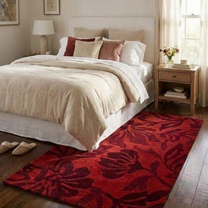 Puede incluir: Un dormitorio con una cama con edred&oacute;n y cojines beige. Una llamativa alfombra roja con un dise&ntilde;o floral est&aacute; sobre el suelo de madera. Tambi&eacute;n se ve una mesita de noche con una l&aacute;mpara y flores.