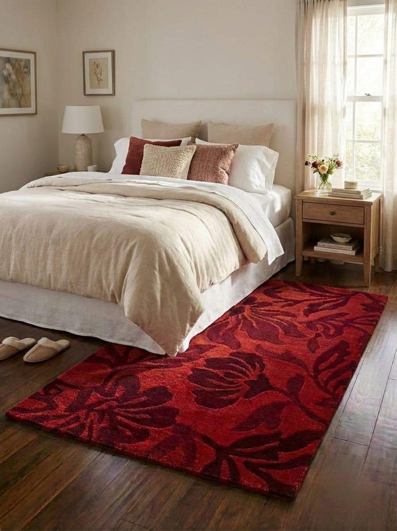 Puede incluir: Un dormitorio con una cama con edred&oacute;n y cojines beige. Una llamativa alfombra roja con un dise&ntilde;o floral est&aacute; sobre el suelo de madera. Tambi&eacute;n se ve una mesita de noche con una l&aacute;mpara y flores.