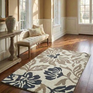 Puede incluir: Una alfombra rectangular con un dise&ntilde;o floral en tonos crema, marr&oacute;n y azul marino. La alfombra presenta grandes dise&ntilde;os estilizados de flores y hojas. La alfombra est&aacute; colocada sobre un suelo de madera en una habitaci&oacute;n luminosa.
