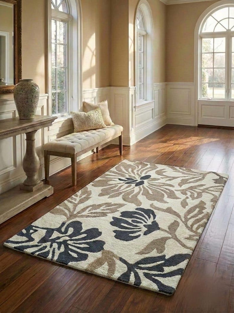 Puede incluir: Una alfombra rectangular con un dise&ntilde;o floral en tonos crema, marr&oacute;n y azul marino. La alfombra presenta grandes dise&ntilde;os estilizados de flores y hojas. La alfombra est&aacute; colocada sobre un suelo de madera en una habitaci&oacute;n luminosa.