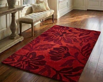 Puede incluir: Una alfombra rectangular roja con un estampado floral en diferentes tonos de rojo. La alfombra está colocada sobre un suelo de madera y el diseño floral es prominente. La alfombra añade un elemento decorativo a la habitación.