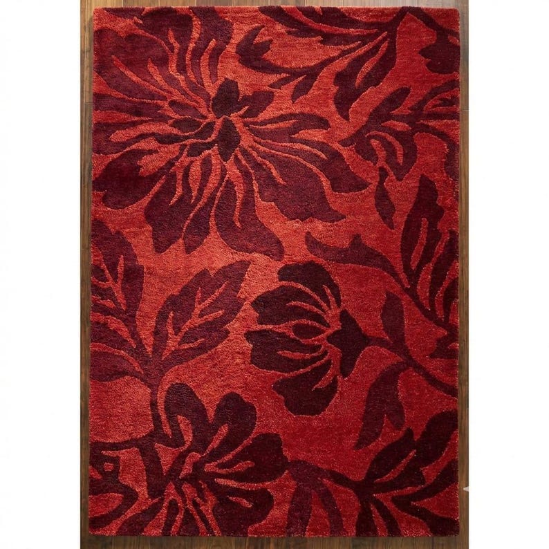 Puede incluir: Una alfombra rectangular roja con un estampado floral en un tono m&aacute;s oscuro de rojo. La alfombra presenta grandes flores y hojas estilizadas, creando un dise&ntilde;o audaz y decorativo. La alfombra est&aacute; colocada sobre un suelo de madera.