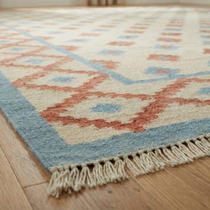 Könnte beinhalten: Ein rechteckiger Teppich mit cremefarbenem Hintergrund und einem sich wiederholenden Muster aus roten und blauen geometrischen Formen. Der Teppich hat einen blauen Rand und Fransen an einer Kante. Er liegt auf einem Holzboden.