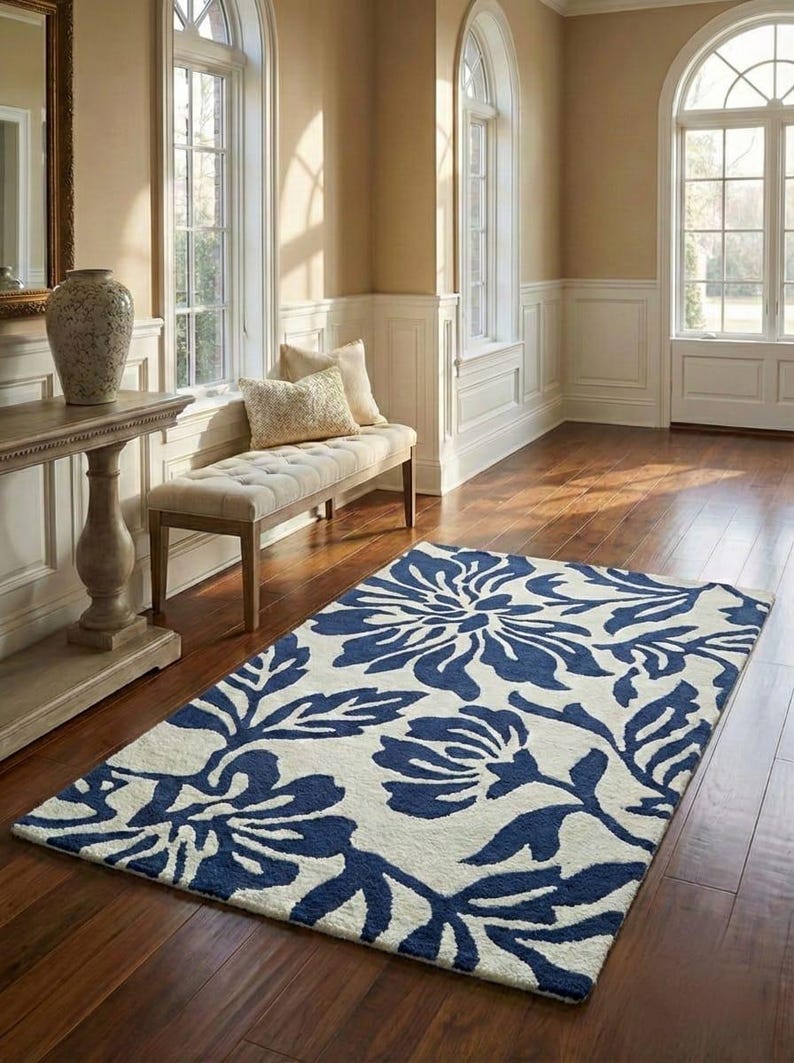 Puede incluir: Una alfombra rectangular con un dise&ntilde;o floral en azul marino y crema. La alfombra est&aacute; colocada sobre un suelo de madera en una habitaci&oacute;n con luz natural y un banco. El patr&oacute;n de la alfombra presenta grandes flores y hojas estilizadas.
