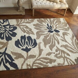 Puede incluir: Una alfombra rectangular con un dise&ntilde;o floral. La alfombra presenta grandes flores en tonos azul marino, marr&oacute;n y beige. Est&aacute; colocada sobre un suelo de madera oscura, con un banco y una mesa auxiliar en el fondo.
