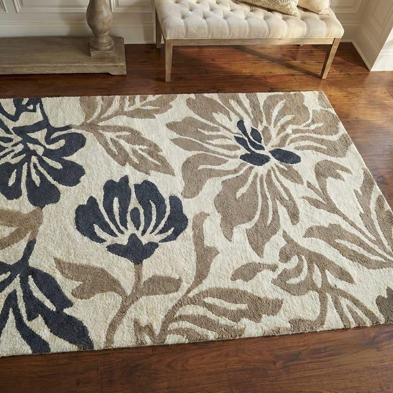 Puede incluir: Una alfombra rectangular con un dise&ntilde;o floral. La alfombra presenta grandes flores en tonos azul marino, marr&oacute;n y beige. Est&aacute; colocada sobre un suelo de madera oscura, con un banco y una mesa auxiliar en el fondo.