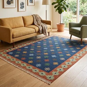 Könnte beinhalten: Ein rechteckiger Teppich mit tiefblauem Zentrum und einem sich wiederholenden Muster aus kleinen, bunten geometrischen Kreuzsymbolen. Der Teppich hat einen breiten Rand in Rostorange mit einem hellgrünen Rautenkettenmotiv und weißen Fransen.