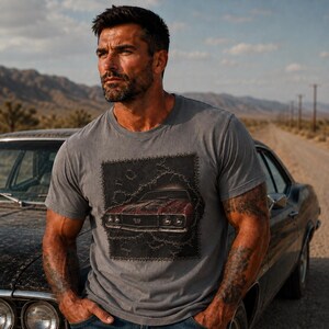 Puede incluir: Una camiseta gris con un gráfico desgastado de un coche rojo clásico con el texto "SS". La camiseta la lleva una persona de pie junto a un coche antiguo en una carretera desértica.