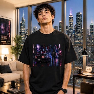May include: Black t-shirt featuring a vibrant graphic of a car in a neon-lit cityscape. The design includes Japanese text and a futuristic aesthetic. The shirt is worn by a person in front of a city skyline.