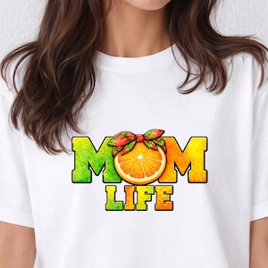 Puede incluir: Camiseta blanca con un diseño gráfico colorido. El diseño incluye las palabras "MOM LIFE" en letras de bloque verdes y naranjas, con una rodaja de naranja y un lazo en el centro. La camiseta es de estilo casual.