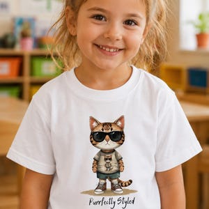Puede incluir: Una camiseta blanca con el gráfico de un gato atigrado marrón que lleva gafas de sol negras, una camiseta beige, pantalones cortos oscuros y zapatillas. El gato está sobre una mancha beige. Debajo, el texto Purrfectly Styled en negro.