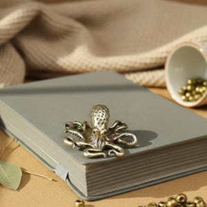 May include: A detailed brass octopus figurine rests on a grey notebook. The octopus has textured details and is positioned on the upper part of the notebook. The notebook is closed and has a grey elastic band. Small gold beads are scattered around the notebook.