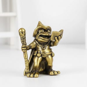 May include: A bronze-colored frog figurine in a wizard's hat and cape, holding a staff and a book. The frog has a detailed face and is wearing a necklace and belt. The figurine is standing upright.