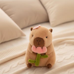 May include: A soft toy capybara in brown tones, clutching a pink flower with a green stem. A small pink flower adorns its head. The toy is placed on a cream-coloured surface.