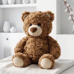 May include: A plush brown teddy bear with a light brown muzzle and paw pads sits on a white blanket. The bear has a friendly expression with black eyes and a stitched smile. The fur is soft and textured.