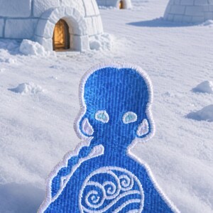 Puede incluir: Un parche bordado azul que presenta una figura estilizada con un borde blanco. La figura tiene un emblema circular en el pecho que muestra un diseño de onda de agua blanca. El parche tiene forma de persona con una cabeza redondeada.