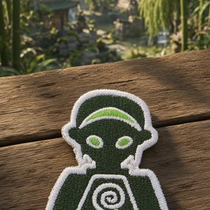 Puede incluir: Un parche bordado que presenta una figura estilizada de color verde oscuro con una diadema y detalles en los ojos de color verde claro. La figura tiene un borde blanco y muestra un símbolo de espiral blanco en el pecho sobre dos barras.
