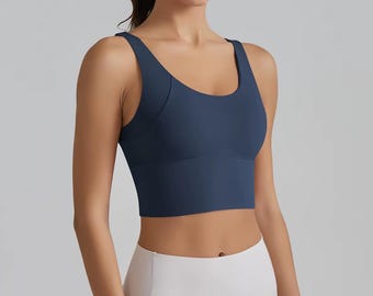 May include: A navy blue sports bra with a scoop neckline. The bra has a wide band under the bust and thick straps. The model is wearing white leggings. The sports bra is designed for athletic activities.