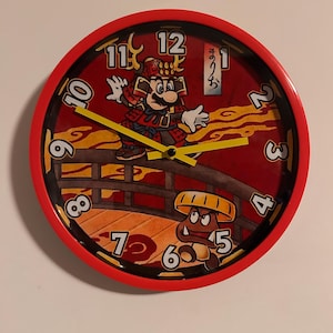 Op de afbeelding: Een rode wandklok met een cartoonafbeelding van Mario in samoerai-outfit op een brug, met een Goomba-personage eronder. De klok heeft een rode wijzerplaat met gele wijzers en witte cijfers.