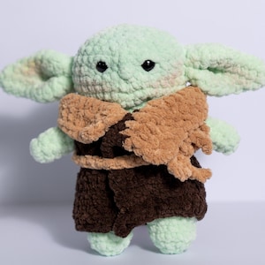 Puede incluir: Mu&ntilde;eco de peluche tejido a ganchillo de un personaje con orejas grandes, piel verde y ojos negros. El mu&ntilde;eco lleva una capa marr&oacute;n y un atuendo marr&oacute;n oscuro. El juguete est&aacute; hecho a mano.
