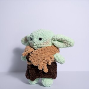 Puede incluir: Mu&ntilde;eco de ganchillo de un personaje con orejas grandes, piel verde y una capa marr&oacute;n. El mu&ntilde;eco tiene ojos negros y viste un atuendo marr&oacute;n. El mu&ntilde;eco est&aacute; hecho de hilo suave y texturizado y est&aacute; de pie.