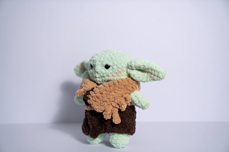 Puede incluir: Mu&ntilde;eco de ganchillo de un personaje con orejas grandes, piel verde y una capa marr&oacute;n. El mu&ntilde;eco tiene ojos negros y viste un atuendo marr&oacute;n. El mu&ntilde;eco est&aacute; hecho de hilo suave y texturizado y est&aacute; de pie.