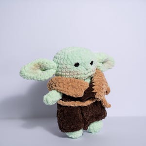 Puede incluir: Mu&ntilde;eco de ganchillo hecho a mano que se asemeja a un personaje popular. El mu&ntilde;eco tiene una cabeza verde claro y orejas grandes, con un abrigo y pantalones cortos marrones. Tiene ojos negros y est&aacute; de pie.