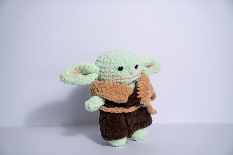 Puede incluir: Mu&ntilde;eco de ganchillo hecho a mano que se asemeja a un personaje popular. El mu&ntilde;eco tiene una cabeza verde claro y orejas grandes, con un abrigo y pantalones cortos marrones. Tiene ojos negros y est&aacute; de pie.