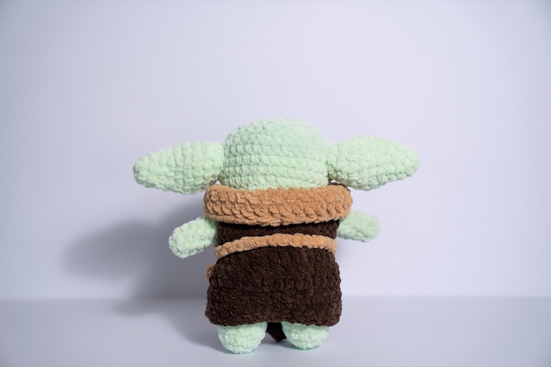 Puede incluir: Mu&ntilde;eca de ganchillo hecha a mano a semejanza de un personaje popular. La mu&ntilde;eca presenta una cabeza y orejas de color verde claro, una t&uacute;nica marr&oacute;n y un cuello color canela. La mu&ntilde;eca est&aacute; hecha de hilo suave y texturizado.