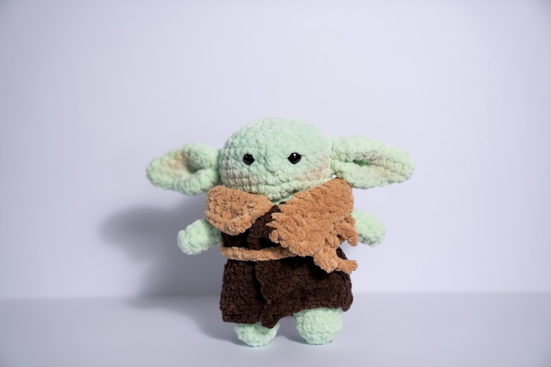 Puede incluir: Mu&ntilde;eco de peluche tejido a ganchillo de un personaje con orejas grandes, piel verde y ojos negros. El mu&ntilde;eco lleva una capa marr&oacute;n y un atuendo marr&oacute;n oscuro. El juguete est&aacute; hecho a mano.