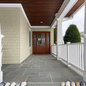 May include: A covered porch with a dark wood ceiling and a gray stone floor. The front door is made of dark wood with glass panels. A row of seashells lines the front step.