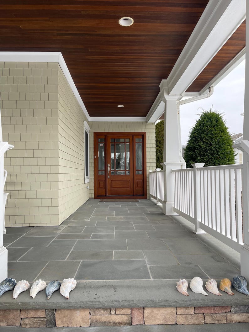 May include: A covered porch with a dark wood ceiling and a gray stone floor. The front door is made of dark wood with glass panels. A row of seashells lines the front step.