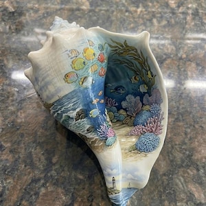 Puede incluir: Una gran caracola que presenta una escena detallada pintada a mano de un arrecife de coral submarino con peces tropicales coloridos, algas y un paisaje de faro costero en las superficies exterior e interior de la concha.