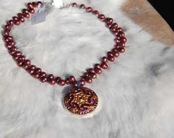 May include: A necklace of burgundy pearls with a decorative pendant. The pendant features a gold filigree design on a purple background. The necklace is displayed on a white, fluffy surface.