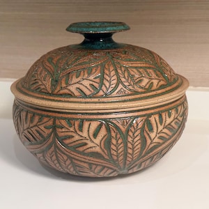May include: A round ceramic lidded jar featuring a tan base with a carved leaf pattern highlighted in dark green glaze. The lid has a matching leaf design and a central knob finished in a deep teal blue glaze, creating a textured appearance.
