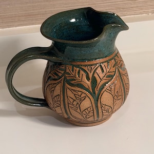 May include: A ceramic pitcher featuring a rounded body with a tan, speckled finish and carved leaf patterns. The upper rim, interior, and handle are glazed in a deep teal blue. The pitcher has a wide spout and a curved handle on the side.