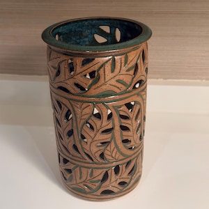 May include: A cylindrical ceramic vase featuring a speckled tan finish with intricate cut-out leaf patterns. The interior and the cut-out edges are glazed in a deep teal color, creating a contrast against the textured tan exterior surface.