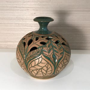May include: A round ceramic vase with a narrow neck and a flared rim. The body features a carved leaf pattern with a tan speckled finish and deep green glaze accents. The upper portion of the vase includes cut-out sections between the leaves.