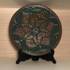 May include: A round ceramic plate featuring a deep teal and brown glaze with a raised, symmetrical floral pattern radiating from the center. The plate is displayed on a black plastic stand, showcasing intricate leaf-like textures and geometric lines.