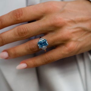 Op de afbeelding: Een ring met een grote, rechthoekige, radiant-geslepen blauwe edelsteen in een zilverkleurige metalen band. De centrale blauwe steen wordt aan weerszijden geflankeerd door twee kleinere, heldere, driehoekig geslepen stenen.
