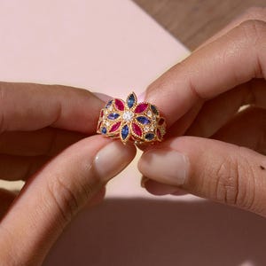May include: A gold ring with a floral design, featuring red, blue, and white gemstones. The ring is held between two hands, showcasing its intricate details and craftsmanship. The gemstones are set in a detailed pattern.