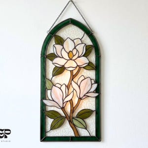 Könnte beinhalten: Buntglasfenster mit Magnolienblüten-Design. Das Kunstwerk zeigt weiße und rosa Blüten mit grünen Blättern und einem braunen Stiel, umrahmt von einem dunkelgrünen Rand. Das Panel wird von einer Kette gehalten.