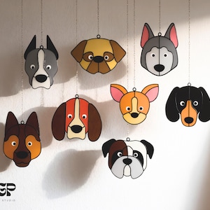 Op de afbeelding: Een verzameling hangende hondenhoofdornamenten in verschillende rassen, waaronder een Husky en een Bulldog. Elk ornament heeft een aparte kleurenpalet en is opgehangen aan een zilveren ketting. Het TGP Design Studio-logo is linksonder zichtbaar.