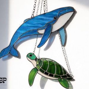 May include: Stained glass whale and sea turtle hanging art. The whale is blue with white accents, and the turtle is green. Both are suspended by chains. The design studio logo is visible in the bottom left corner.