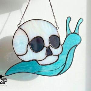 May include: Stained glass art piece depicting a snail with a skull-shaped shell. The shell is white with blue marbling, and the snail's body is a vibrant turquoise. The piece is suspended by a chain.