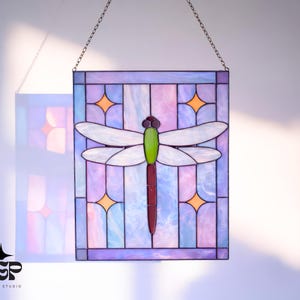 Pode incluir: Painel de vitral quadrado com um desenho de libélula. A libélula tem asas brancas, cabeça verde e corpo bordô. O fundo é uma mistura de roxo, rosa e azul com estrelas laranja. Pendurado por uma corrente.