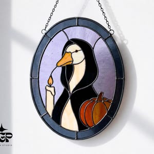 May include: Oval stained glass window depicting a goose in a black cloak, holding a candle and a pumpkin. The window has a black frame and is suspended by a chain. The goose has a yellow beak against a purple background.