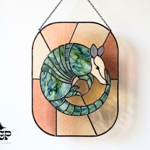 Könnte beinhalten: Buntglasfenster mit einem eingerollten Gürteltier in Grün, Blau und Weiß, vor einem Hintergrund aus Pfirsich- und Beigesegmenten. Das Kunstwerk ist schwarz umrahmt und an einer Kette aufgehängt.