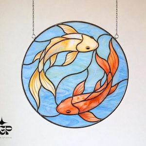 May include: Circular stained glass artwork depicting two koi carp in yellow and orange hues, set against a blue water backdrop. The design is framed with a black border and hangs from a chain.